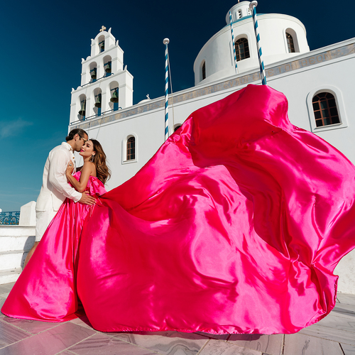 Couple beside a white bell tower in Santorini, the woman in a bright pink ruffled flying dress