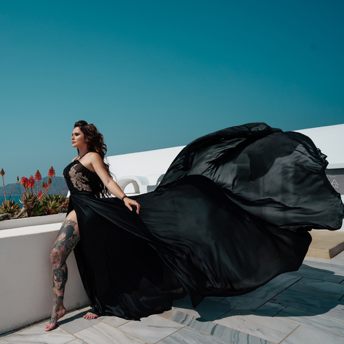 Woman in a black flying dress on a white terrace, the long train blowing in the wind in Santorini