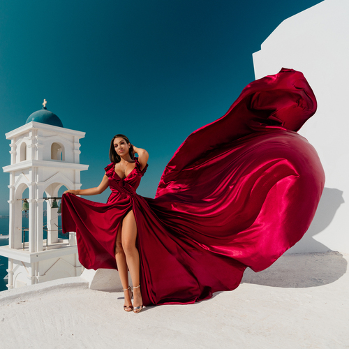 Woman in a burgundy flying dress with a high slit beside a white bell tower in Santorini