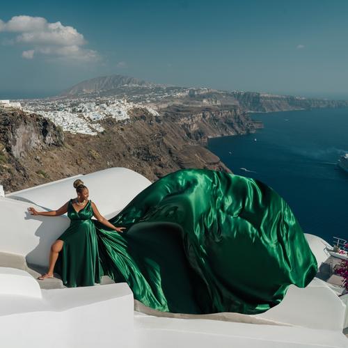 Woman in a deep green flying dress seated on a white wall above the Santorini caldera