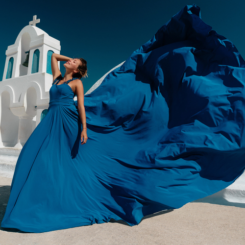 Woman in a blue flying dress beside a white bell tower, the long train filling the frame, Santorini