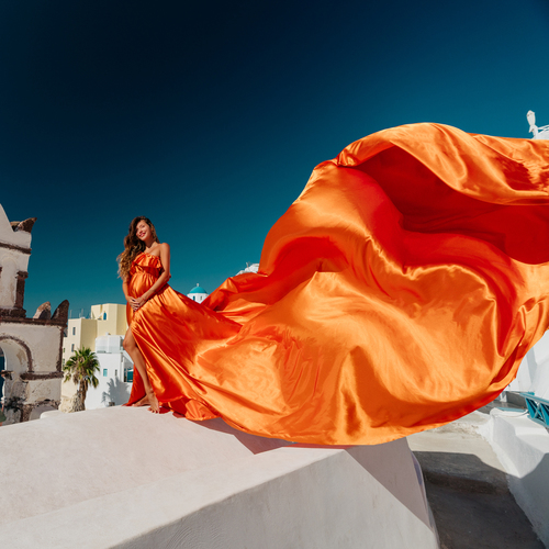 Woman in an orange ruffled flying dress, the train billowing against a deep blue sky in Santorini