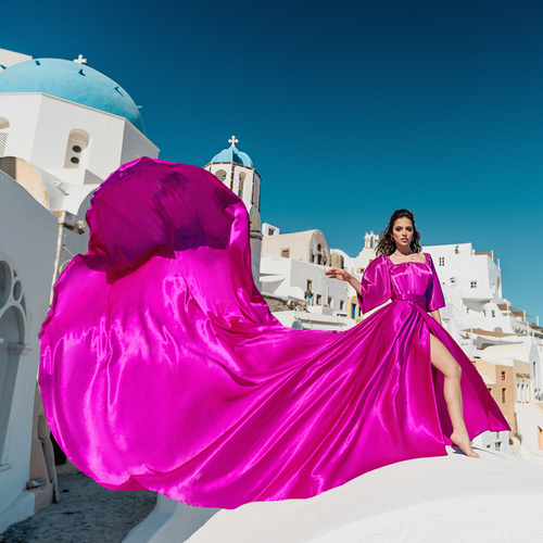 Woman in a fuchsia flying dress with sleeves and a flowing train, whitewashed Santorini village behind