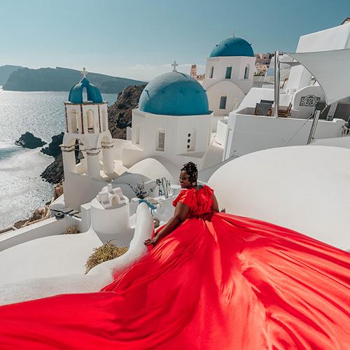 Woman in a red ruffled flying dress seated on a white terrace with blue domes behind, Santorini