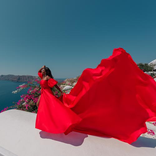 Woman in a red flying dress with sleeves, the train blowing across a white rooftop above the sea in Santorini