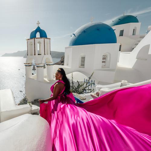 Woman in a hot pink flying dress seated in front of blue-domed churches in Santorini